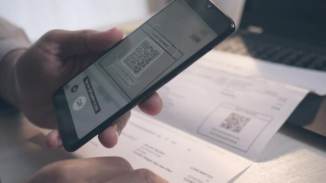 Handheld shot of a male hand holding smartphone, scanning a QR code on the invoice in front of him, and making the payment transaction