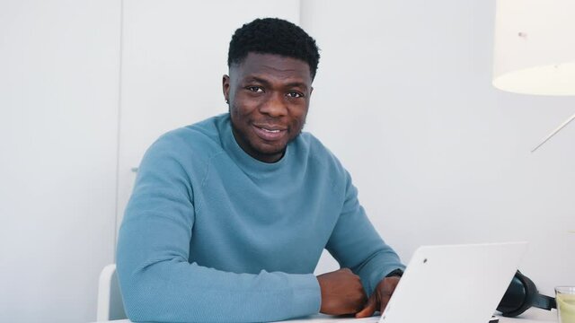 African American black man sitting at the table in front of a laptop working. Man looking at the camera and smiling while workingon the laptop. Headphones and a table lamp can be seen. 