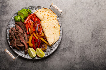 Traditional Mexican dish Beef fajita served with tortillas and ripe avocado on rustic metal plate and stone background from above, American Mexican food healthy meal with space for text
