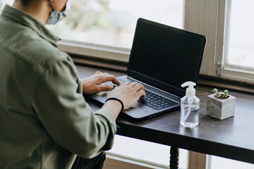 young woman sitting in a mask at coffee shop working on laptop, using disinfectant