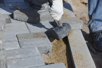 A worker knocks on the paving slabs with a construction hammer, aligns small concrete blocks laid in the sand.