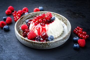 Modern style traditional blancmange almond pudding with wild berry coulis served as close-up in a Nordic design plate on black background