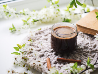 Obraz na płótnie Canvas Spring still life on a white windowsill, a cup of aromatic espresso coffee, good morning concept. Blooming branches of white cherry. Scandinavian style, knitted stand