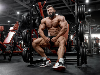Naklejka na ściany i meble A brutal man with a beard is sitting on the bench of the simulator in the gym. A muscular bodybuilder in perfect athletic shape rests between sets in training
