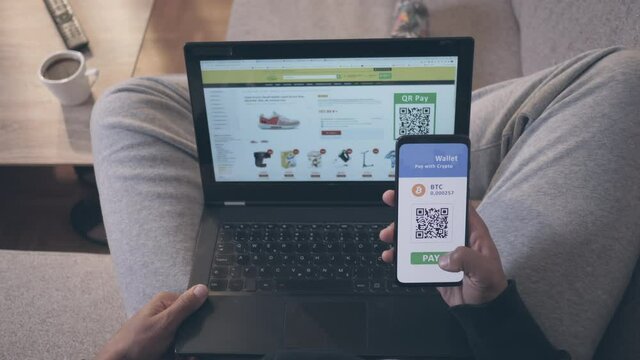 POV Shot Of An African-American Man Sitting In The Living Room, Holding A Laptop In His Lap And Smartphone In Right Hand, Shopping Online With QR Scan On Mobile Crypto Wallet App