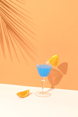 Blue cocktail drink decorated with a slice of orange. In the background shade of tropical palm leaves on a combination of light brown and cream background. Summer vibes party concept.