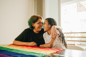 Pareja de jovenes lesbianas con bandera LGTBQ se abraza en la mesa del comedor de su casa