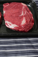 Fresh raw rib eye steak on a plastic tray on a black and white stripe butchers apron. Meat industry product. High quality beef with excellent marbling.