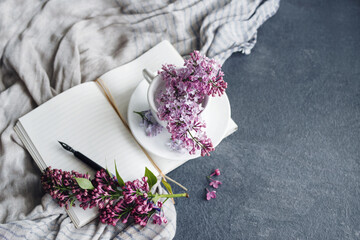 Spring romantic background with lilac flowers in a white cup, with a open white notebook and a pen...