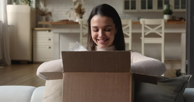 Woman sit on sofa at home open courier parcel, glad customer receive delivered goods check items inside of box feels satisfied. Client of e-commerce, postal shipping, express delivery services concept