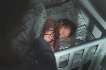 Mother putting her newborn baby two months old to sleep in the cradle bed at night in dark room -...