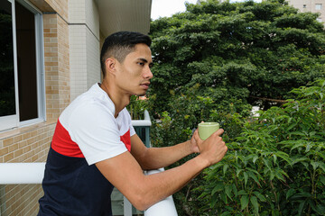 Young Brazilian man holding a cup of coffee, looking ahead, leaning on the balustrade of the...