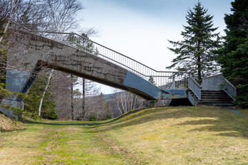 Fototapeta na wymiar A vintage concrete pathway cantilever bridge spanning over a path in a park. The concrete bridge has steps up on both sides with a grey metal handrail. There are trees in the background with blue sky.