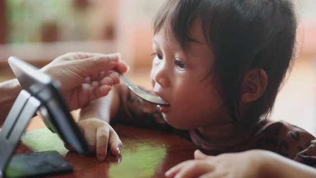 The little cute beautiful girl watched her smartphone with focus and attention while eating the food her mother fed. The concept of children is overly addicted to smartphones and is fed up with food.