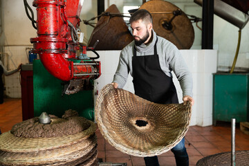 Obraz na płótnie Canvas Focused young man working in handicraft olive oil producing factory, spreading milled olives on fiber disks to press..