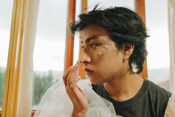 Fototapeta na wymiar Joven queer con maquillaje artístico se observa en el espejo mientras se coge el rostro con la punta de sus dedos 