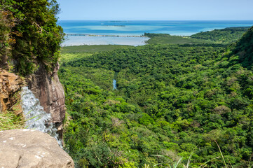 View from the upper base of Pinaisara waterfall, flowing. Sea in blue tones of Okinawa, lush mangrove forest and main road between sea and river in the background. Iriomote Island.