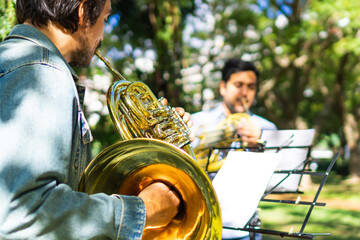 latin man playing a french horn in the park while rehearsing with sheet music, playing in a group....