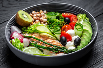 Grilled chicken, avocado, asparagus, chickpeas, broccoli, radish, chicken, cucumber, tomatoes, olives, mozzarella buddha bowl on dark background, top view. Delicious balanced food concept