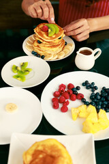 Asian happy skilled pastry housewife in  red apron decorate stack of pancake dessert topping with mixed fruit kiwi raspberry apple from material dish in blurred foreground