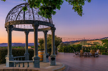 Outdoor al fresco chairs and table on a wooden deck at sunset in the spring with grape vines and hills in the background, Napa Valley, California USA