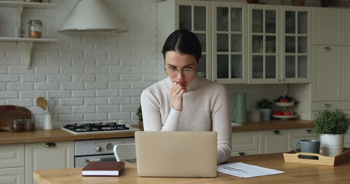 Serious confident concentrated 30s female sit at kitchen table wear eyeglasses working on wireless computer, consider telecommute creative task, texting on modern device, workflow from home concept