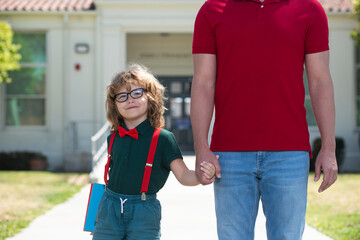 Parent and pupil of primary school go hand in hand. Teacher in t-shirt and cute schoolboy with backpack near school park. Portrait of happy nerd pupil holding teachers hand.