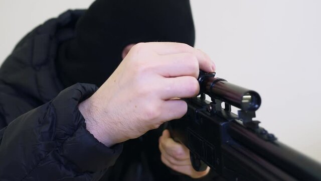A terrorist in a black balaclava adjusts his sniper rifle, makes adjustments, twitches the bolt of the rifle and aims at the victim. Close-up of weapons.