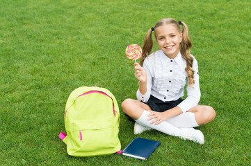 Happy girl child in school uniform hold sweet lollipop sitting on green grass, candy