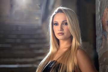 Fototapeta premium Beautiful young long haired blond woman with serious face expression wearing black top and black long pants stand on staircase inside of old rustic house illuminated with ambient light