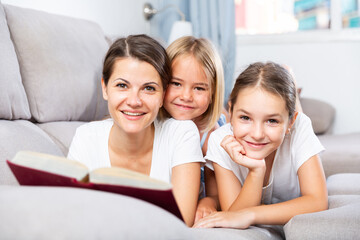 Portrait of happy single mother lying on sofa reading book to her daughters