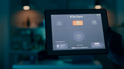 Close up shot of tablet with active smart home application holded by man giving voice command to switch on the lights. Person using high technology software working remote at laptop