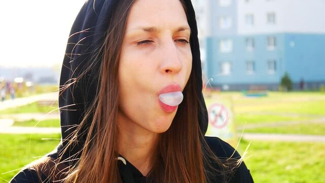 A beautiful girl in a black hood chewing white gum and inflates a bubble against a green lawn background close-up
