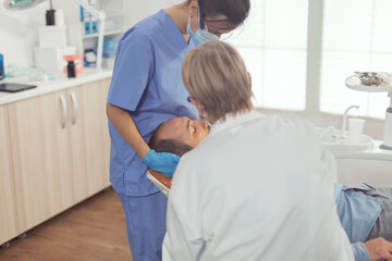 Obraz premium Sick man sitting on dental chair while senior doctor taking care of tooth health. Stomatologist speaking to patient while nurse cleaning mouth during dental examination in stomatology clinic