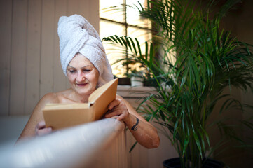 Happy senior woman reading book in bath tub at home.