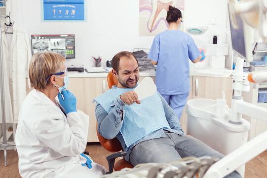 Sick patient checking health teeth analyzing surgery procedure in mirror. Man sitting on dental chair while senior dentist woman explaining stomatological treatment for toothache