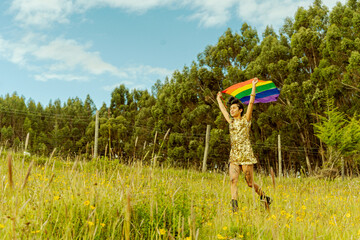 Mujer transgenero corriendo frente al bosque con la bandera LGTBQ en alto  - obrazy, fototapety, plakaty
