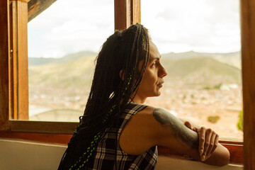 Mujer transgenero tatuada y con trenzas mirando el paisaje a través de su ventana  - obrazy, fototapety, plakaty