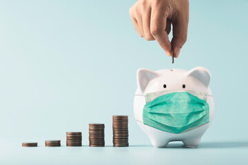 Hand putting coin to white piggy bank saving which wearing surgical mask on blue background and...