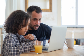 Focused dad and son doing homework using laptop. Curly-haired serious boy and bearded father sitting at table, typing on laptop, looking at screen. Modern technology, education, family concept