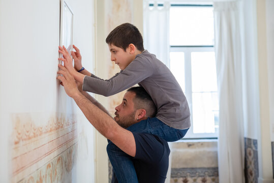Concentrated son and bearded father hanging picture on wall. Caucasian boy sitting on mans neck, looking at picture, decorating apartment during renovation. Side view shot. Family concept.