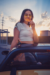 Young female in sunglasses and white t-shirt is eating pizza while posing standing in yellow car...