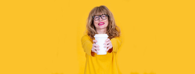 Drink this! adult  woman in spectacles, holds disposable cup of coffee, indicates at beverage, poses against yellow background with free space aside for your promotional content