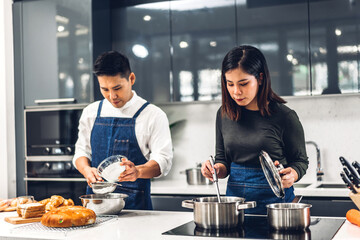 Young asian family couple having fun standing near stove and cooking together.Happy couple looking and smelling tasting fresh delicious from soup in a pot with steam at white interior kitchen