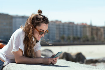 Young happy smiling woman checking her phone at the coastline of a city.