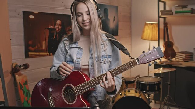 Tilt-up medium slowmo portrait of young smiling female guitarist with long blond hair in vintage denim jacket looking at camera playing acoustic guitar in cozy retro-style music studio