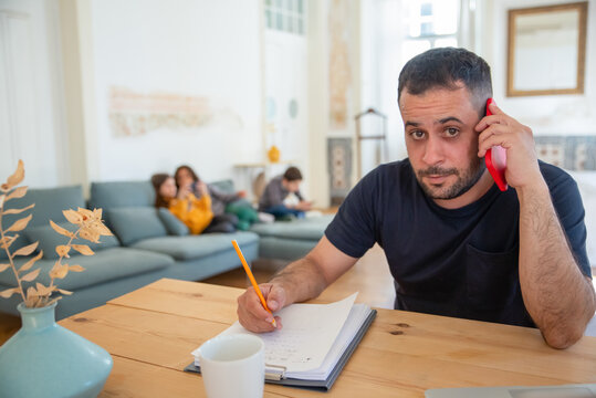 Focused Caucasian Dad Talking On Phone And Looking At Camera. Bearded Man Working At Home, Sitting At Table And Writing Information On Paper. Fatherhood, Work At Home Concept