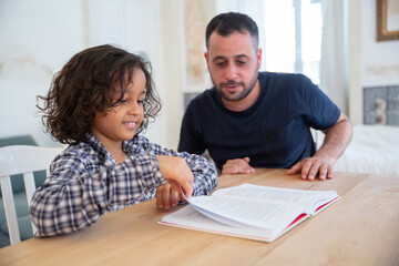 Curly-haired boy sitting with father at table and studying. Cheerful son turning page of textbook...