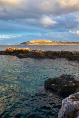 Rocks leading into the ocean and sun hitting a hill in the background on a fall evening in Qawra, Malta.