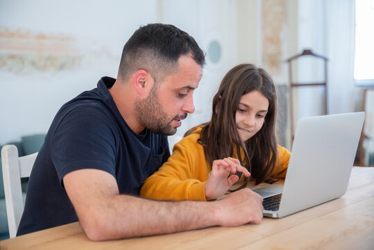 Bearded dad teaching daughter to type on keyboard. Focused brunette girl sitting at table with father, doing homework using laptop. Modern technology, education, family concept